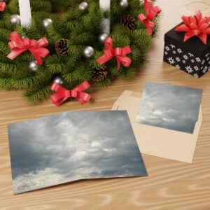 A picture of a few greeting cards on a desk next to an evergreen wreath with candles and ribbons, there is also a small gift box in the arrangement. The greeting card depicts a cloudy, dark grey, sky.