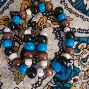 Close-up of a beaded bracelet and earrings featuring turquoise-blue, black, brown wood, and off-white bone beads on a detailed blue and gold paisley background.