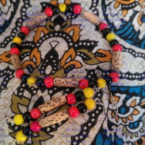 Close-up of a handmade jewelry set featuring red, yellow, and black glass beads combined with light tan carved wooden beads. The matching bracelet and earrings rest on a blue, white, and gold paisley-patterned fabric background.